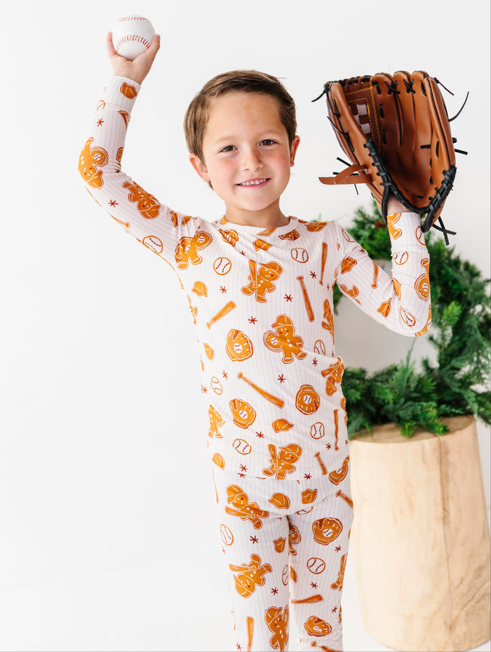 A young boy wearing a white long-sleeve pajama set with an orange baseball-themed print, holding a baseball and glove in front of a white background with a potted plant.
