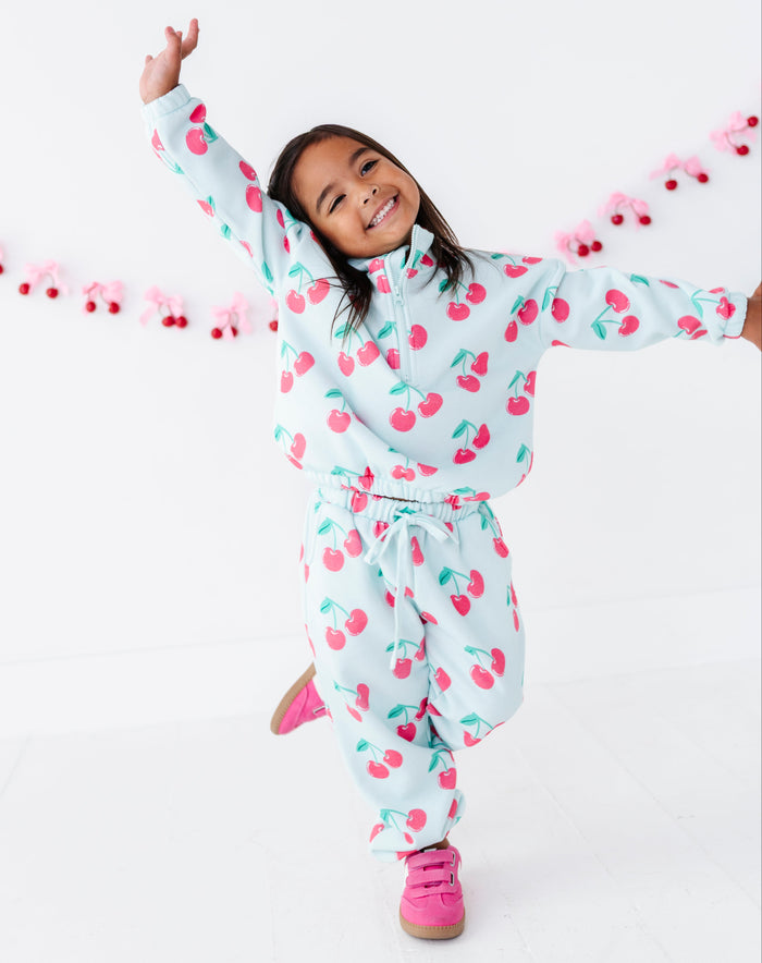 A young girl smiling and posing in cherry-patterned zip-up sweatshirt and matching sweatpants, with bright pink sneakers, against a clean white studio background with a pink cherry garland.
