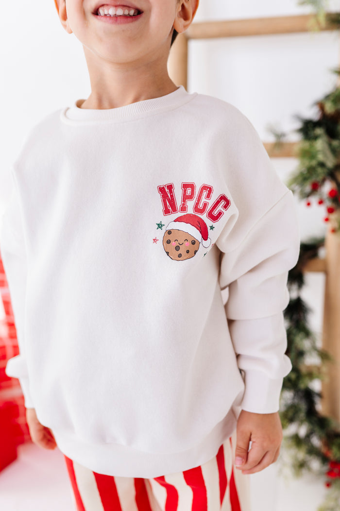 A child wearing a white crewneck sweatshirt with a red and brown 'NPCC' cookie graphic on the chest, paired with red and white striped pants, in a festive holiday setting.