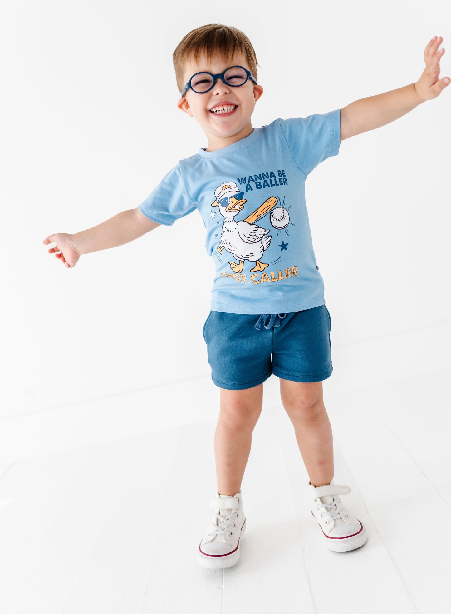 Little boy wearing a baseball tee and dusty blue bamboo cotton shorts standing against a white background