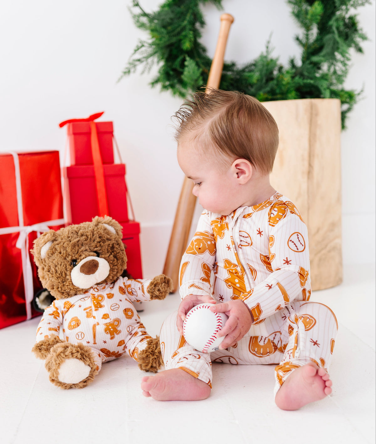 A baby wearing a white long-sleeve pajama romper with an orange holiday print sits on a white floor next to a teddy bear in matching pajamas, holding a white baseball. A green wreath, wooden bat, and red gift boxes are in the background.