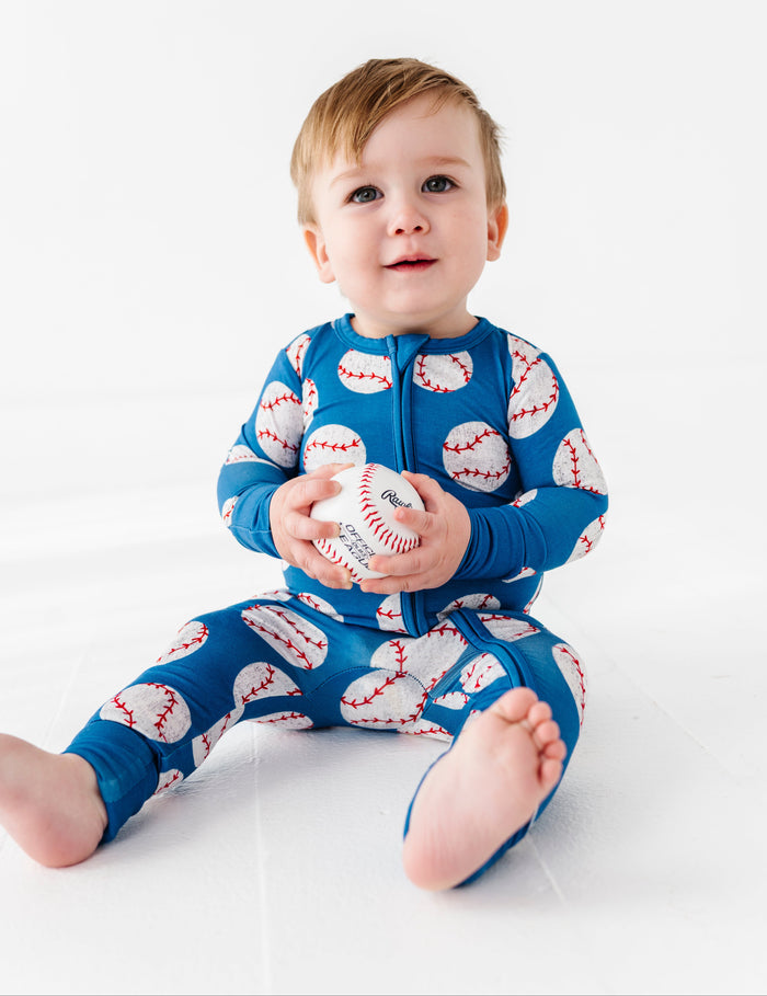 Child wearing a blue bamboo convertible romper with baseball pattern holding a small ball on a white background