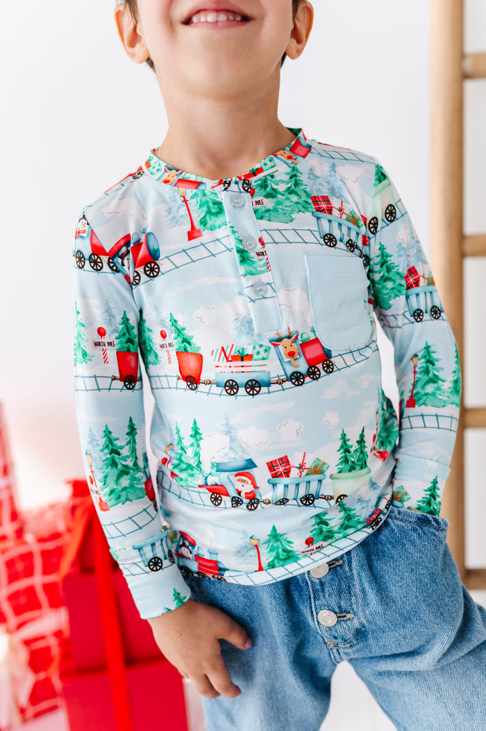 A child wearing a long-sleeve henley shirt with a festive Christmas train pattern, paired with blue jeans. The shirt features a light blue background with red, green, and white holiday illustrations of trains, pine trees, and snowflakes.