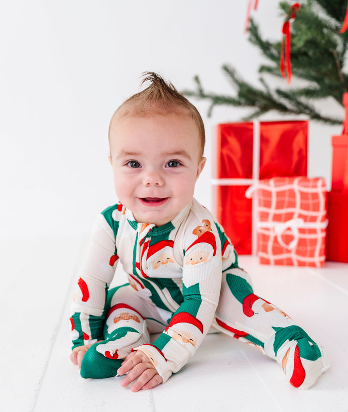 A baby wearing a festive Christmas-themed footed bamboo sleeper sits on a white wooden floor next to a decorated Christmas tree and wrapped presents.