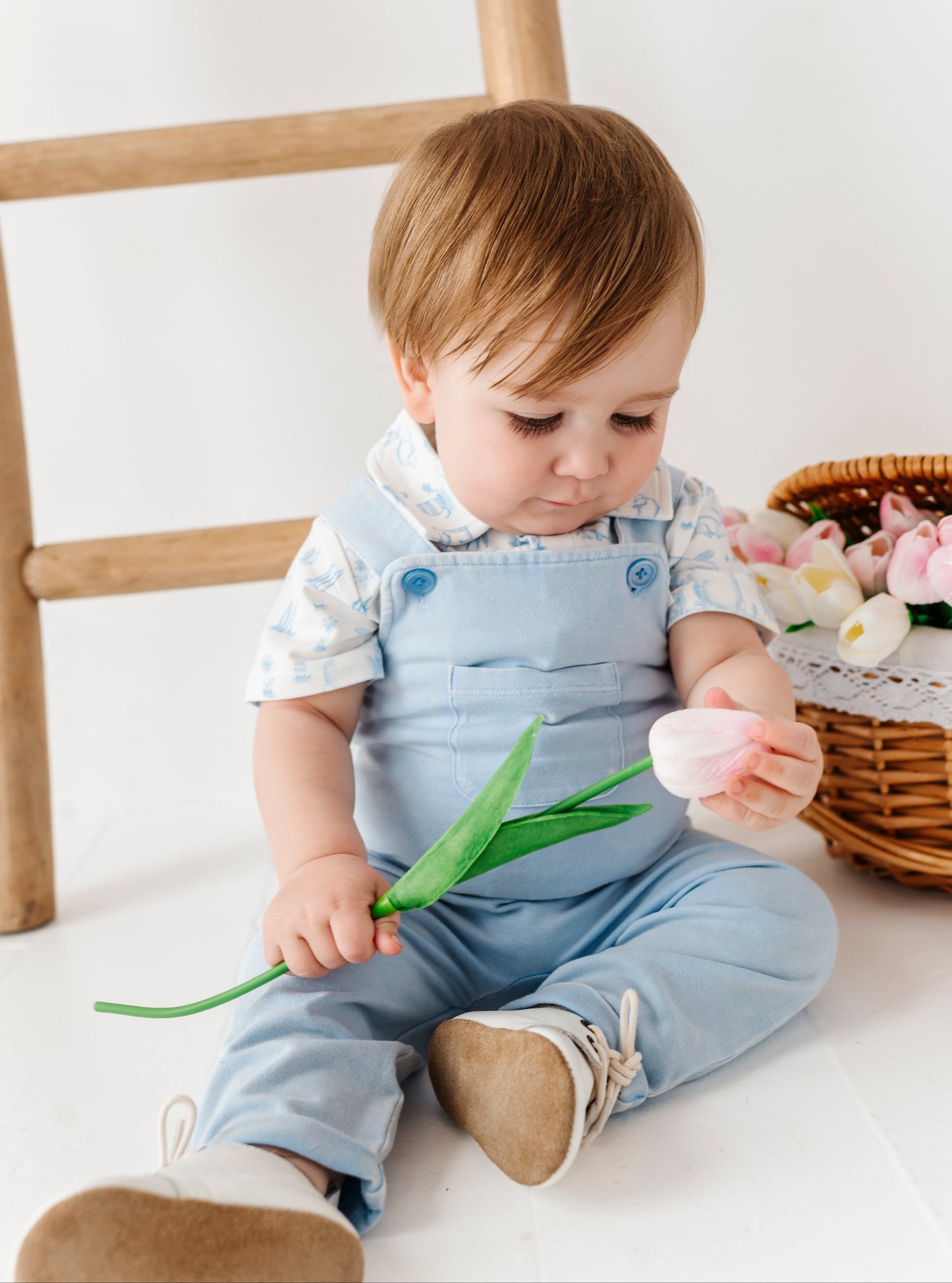 Child in denim overalls holding a tulip and an egg on a white floor with a wooden chair and basket of flowers in the background.