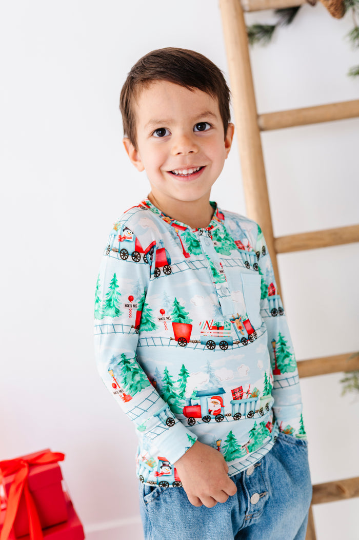 A child wearing a long-sleeve bamboo henley shirt with a festive Christmas train pattern, paired with blue jeans. The shirt features a light blue background with red, green, and white holiday illustrations of trains, pine trees, and snowflakes.
