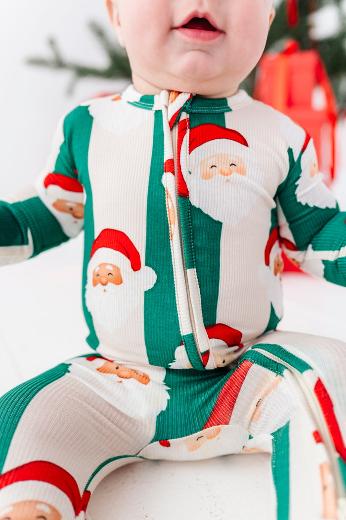 A baby wearing a festive Christmas-themed zip-up bamboo sleeper with a Santa Claus pattern, sitting against a soft white background with blurred holiday decorations.