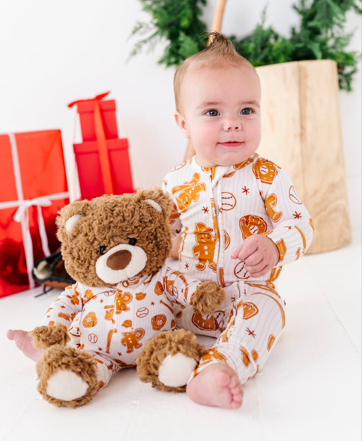 A baby wearing a white and orange holiday-themed footed onesie sits on a white floor next to a brown teddy bear, with red gift boxes and a green wreath in the background.