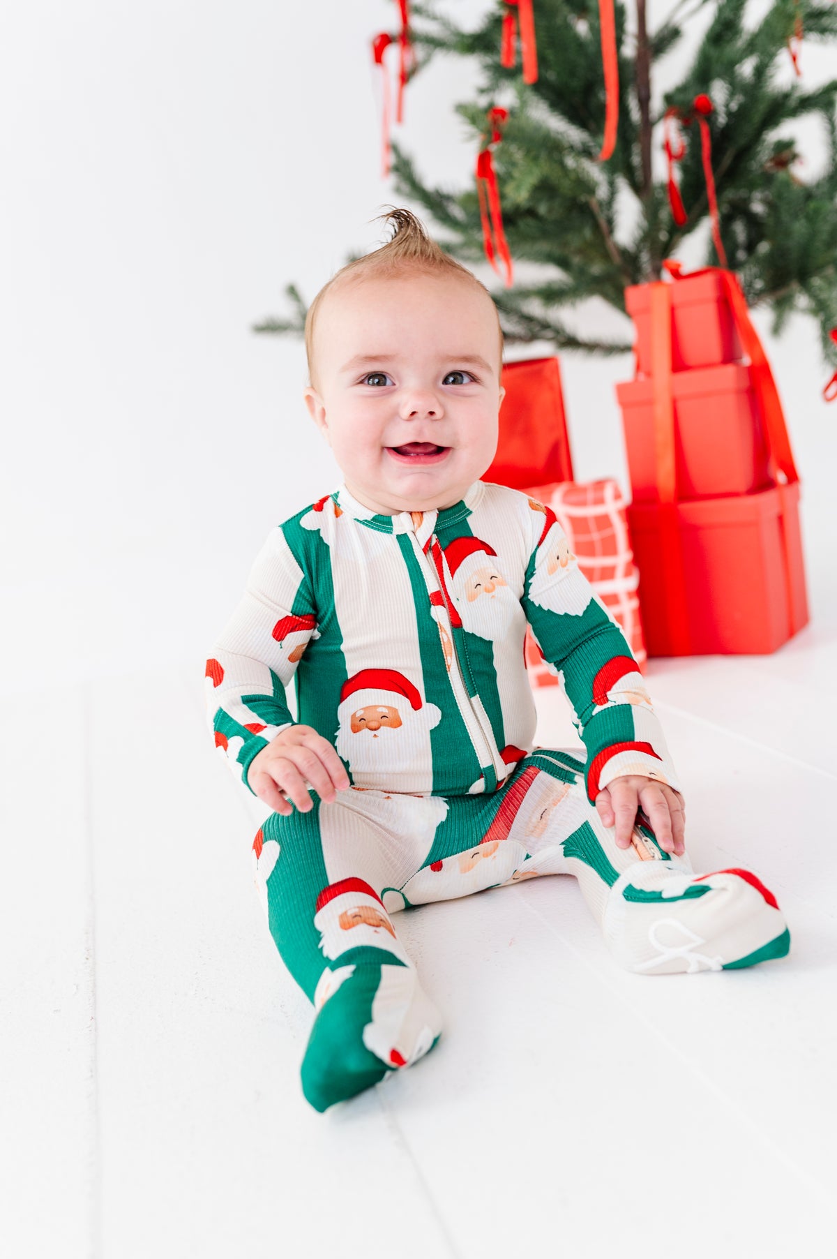 A baby sitting on a white floor wearing a festive green and white Santa Claus print footed bamboo pajama, with a Christmas tree and red wrapped gifts in the background.