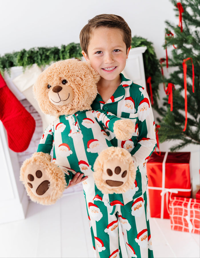A young boy smiling while wearing festive green and white Santa pajamas, holding a large beige teddy bear and matching plush paw slippers in a decorated holiday room with a Christmas tree and wrapped presents.