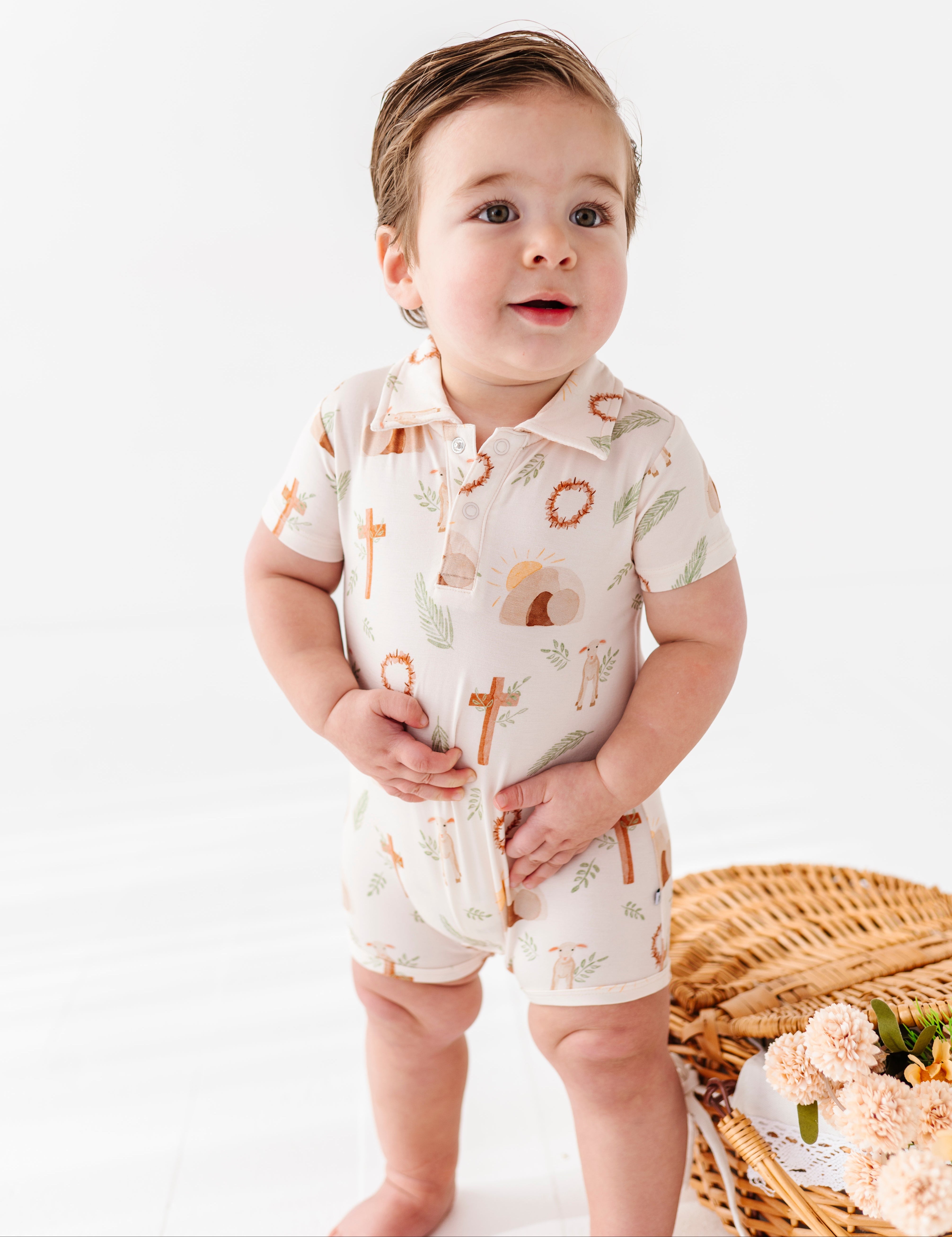 Baby wearing an Easter bamboo romper standing next to a woven basket with flowers on a white background