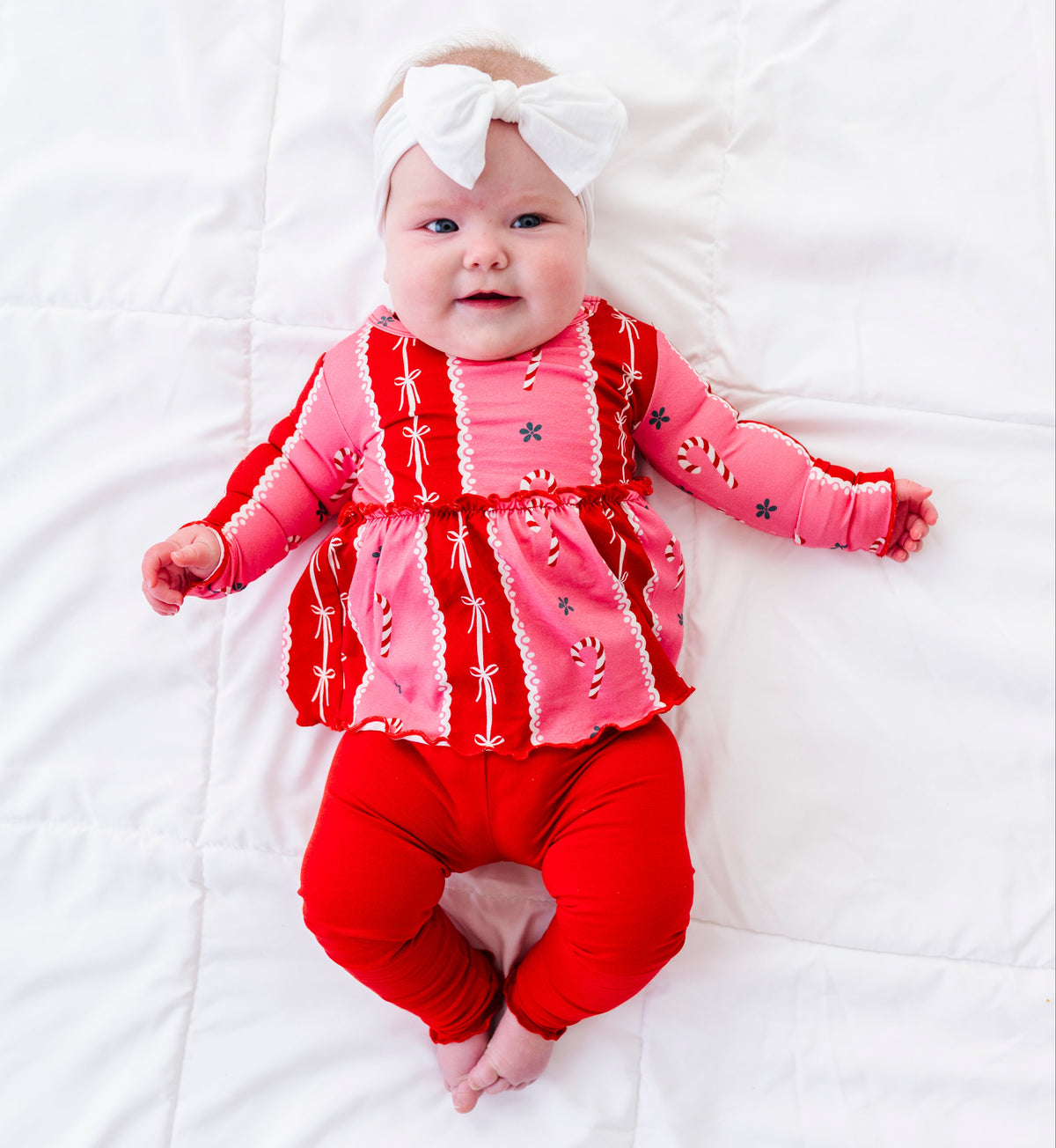 A baby lying on a white quilted blanket, wearing a pink and red patterned long-sleeve bamboo top, red pants, and a white bow headband.