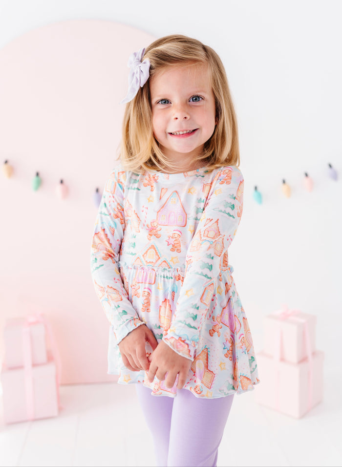 A young girl smiling and wearing a gingerbread village-themed bamboo peplum top with soft purple leggings.