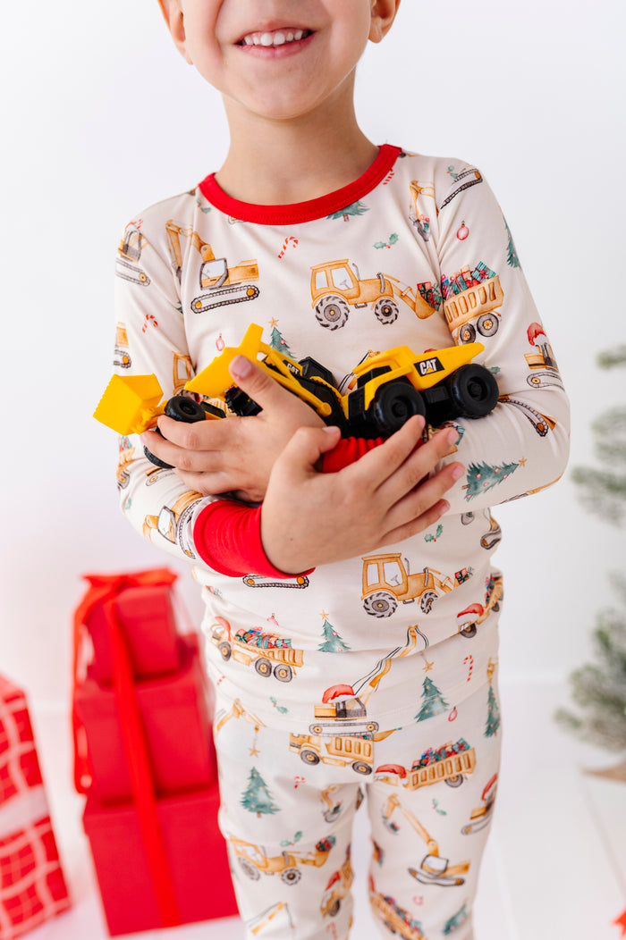 A child wearing cream-colored long-sleeve bamboo pajamas with a construction vehicle print, holding two yellow toy dump trucks. The pajamas feature red trim at the collar and cuffs.