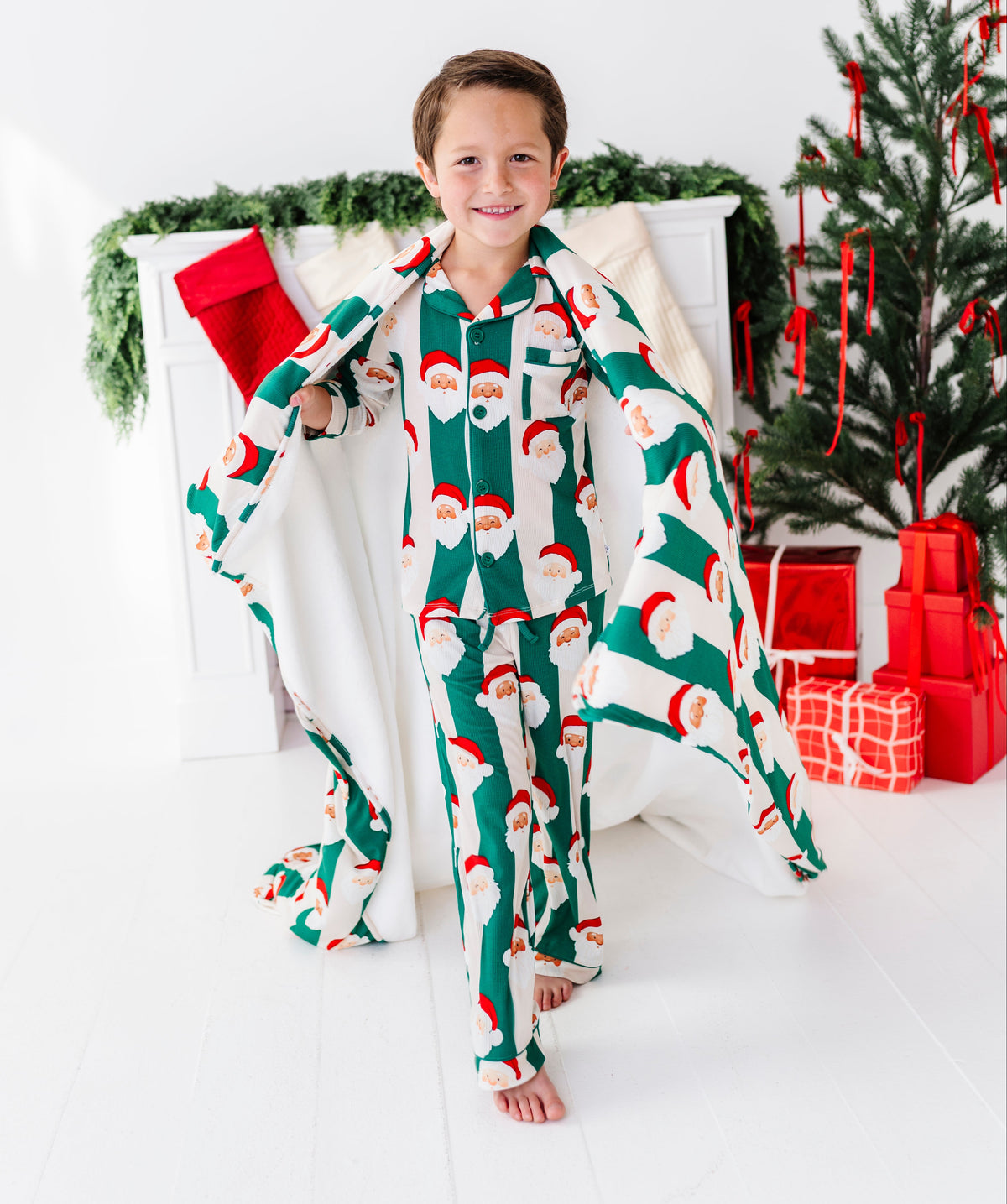 A young boy stands barefoot in a holiday-themed room, wearing a green and white Santa Claus patterned pajama set with a matching cape. A Christmas tree, stockings, and wrapped gifts are visible in the background.