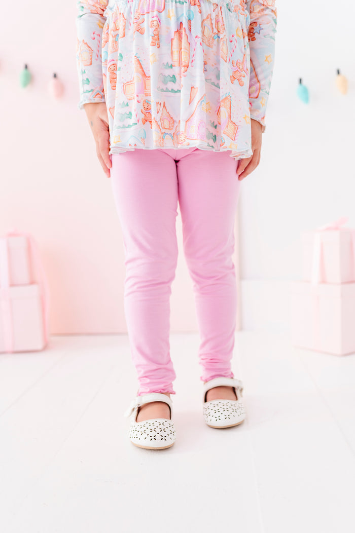 A child wearing pink bamboo leggings, a pastel multicolor patterned long-sleeve peplum top, and white embellished Mary Jane shoes, standing against a soft pink studio background.
