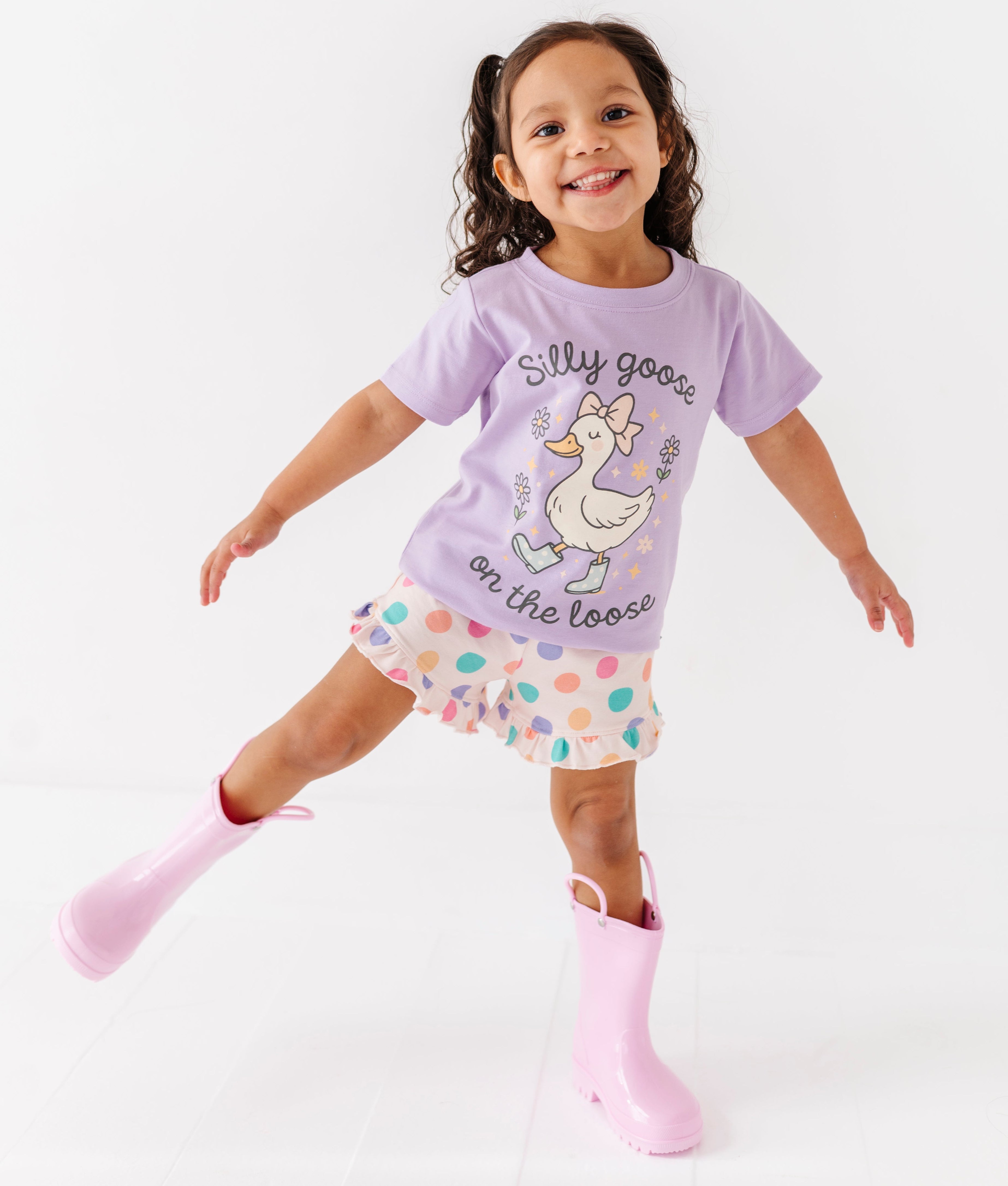 Child wearing a purple shirt with a cartoon goose and silly goose on the loose text, polka dot shorts, and pink rain boots on a white background