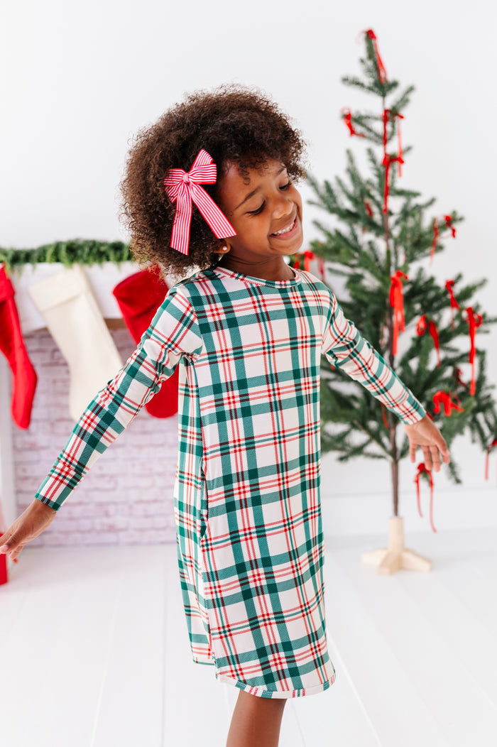 A little girl wearing a bamboo nightgown in a classic Christmas plaid print, standing in front of a fireplace with stockings and a decorated Christmas tree