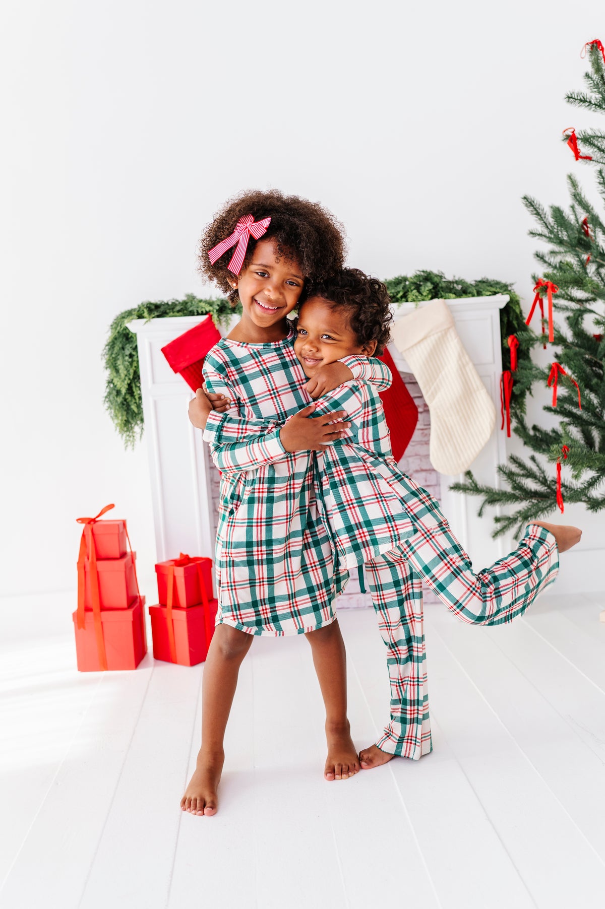A little girl wearing a bamboo nightgown in a classic Christmas plaid print, hugging her little brother who is wearing a matching classic Christmas plaid lounge set