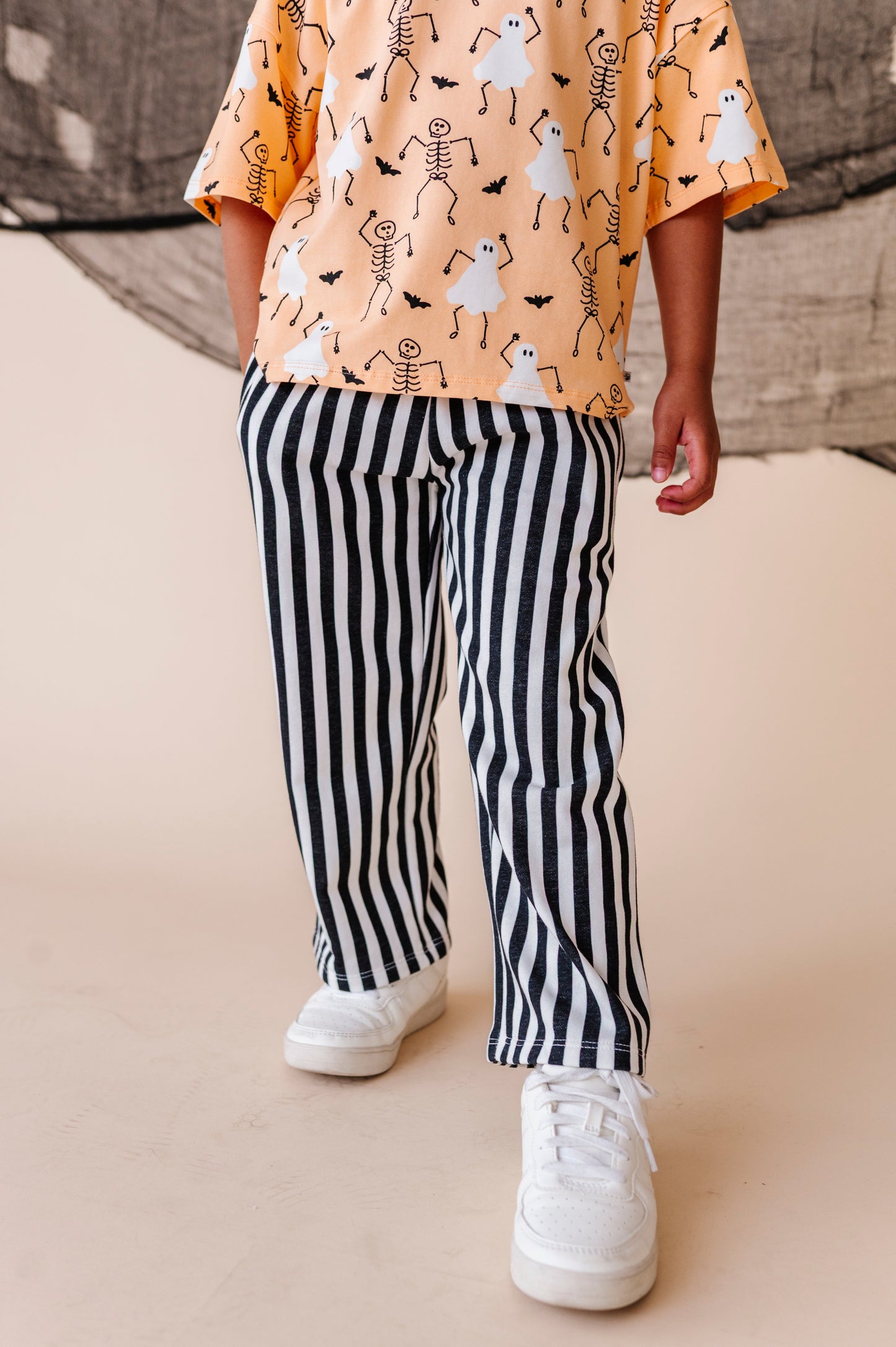 Child wearing a Halloween-themed shirt with skeletons and ghosts, black and white striped pants, and white sneakers on a beige background.