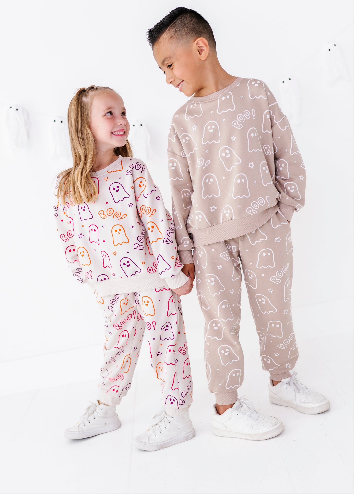 Two children modeling matching ghost-print sweatshirt and jogger sets in a bright white studio. The girl wears a white set with pink, orange, and purple ghosts, and the boy wears a beige set with white ghosts.
