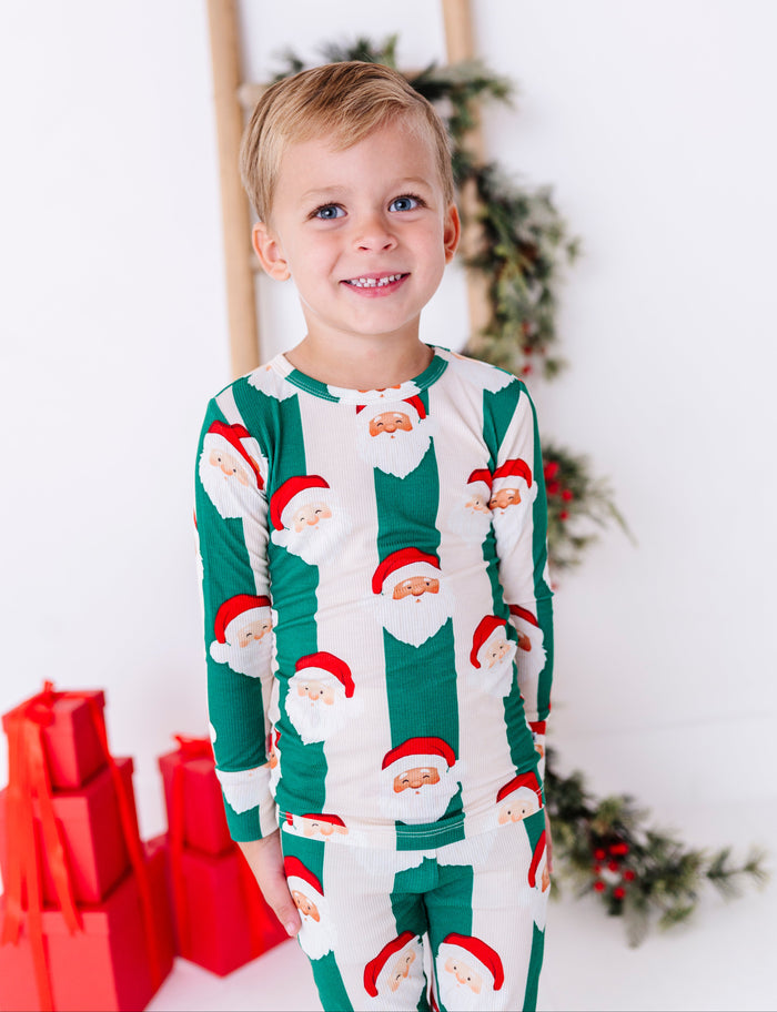 A young blonde boy wearing green-and-beige striped bamboo pajamas with Santa faces all over them. The super soft bamboo pajamas are ribbed and made to fit snugly for safety standards