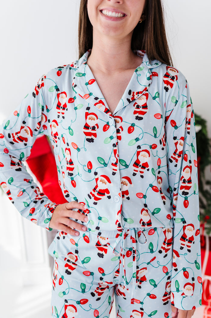A woman wearing a light blue long-sleeve bamboo pajama set with a festive Christmas lights and Santa Claus print, holding a red gift bag.
