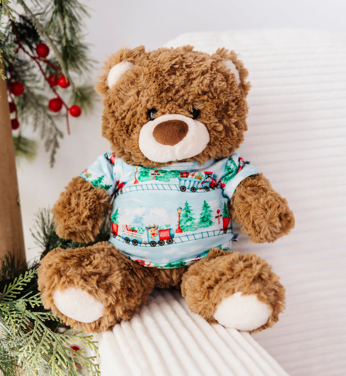 A brown plush teddy bear wearing a light blue Christmas-themed bamboo t-shirt with a train pattern, sitting on a white ribbed chair next to a decorated Christmas tree branch with red berries.