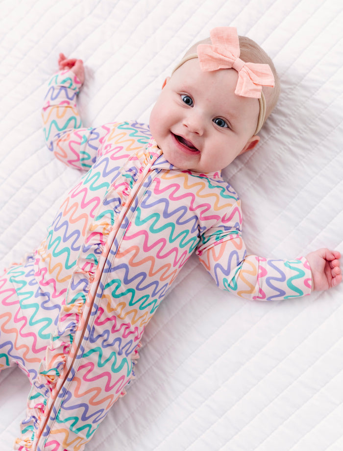 Baby wearing a colorful bamboo zip romper with a pink bow on a white quilted surface