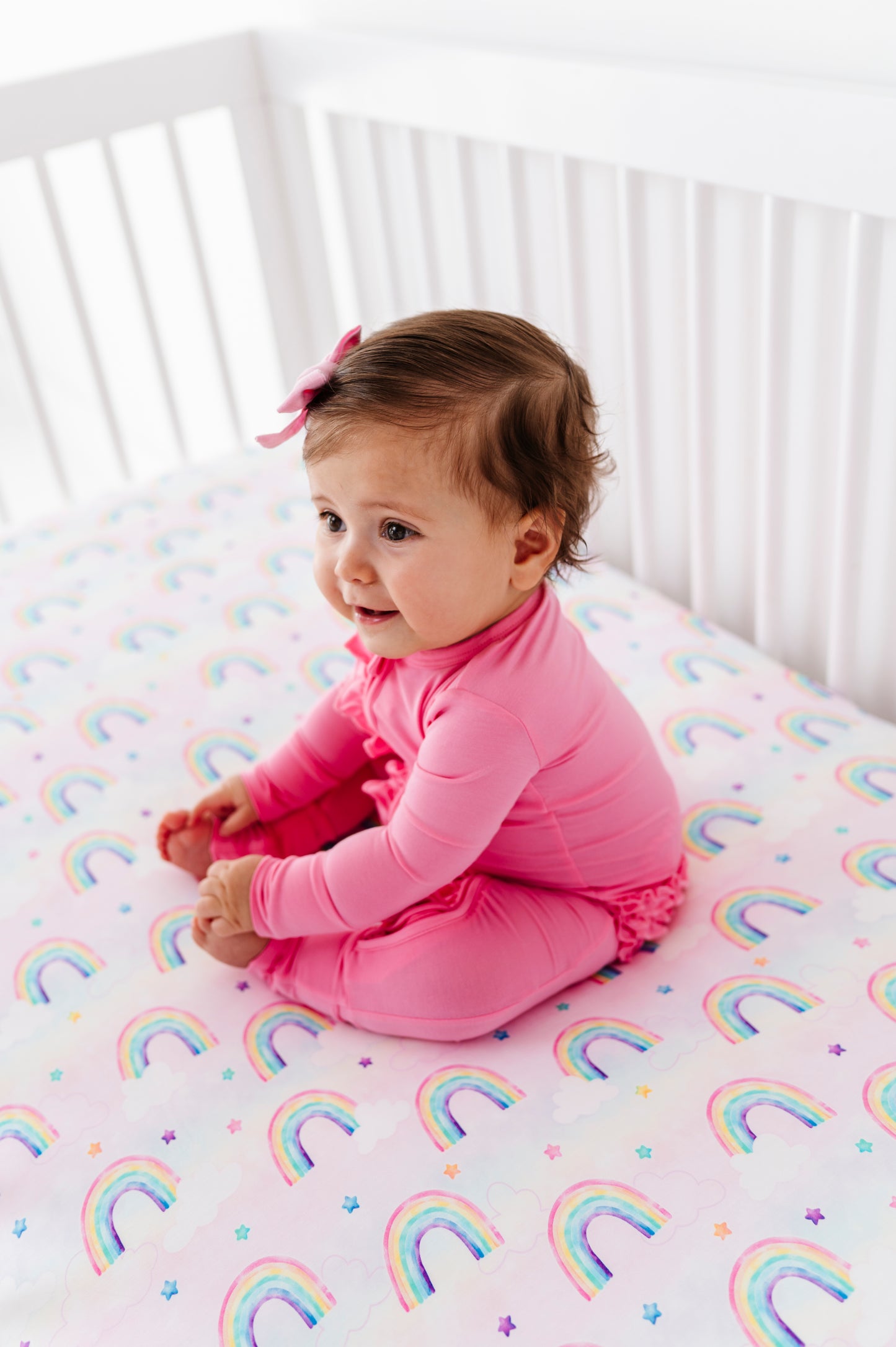 Baby wearing a bamboo convertible romper and sitting on a crib with a rainbow-patterned sheet