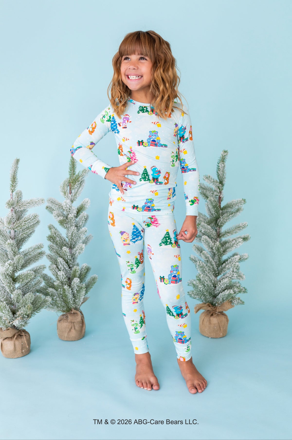A young girl stands barefoot against a light blue studio backdrop, wearing a matching long-sleeve pajama top and leggings set with a colorful Christmas tree and snowman print on a white base. Small potted frosted trees are placed around her, and a copyright notice for ABC-Care Bears LLC appears at the bottom.