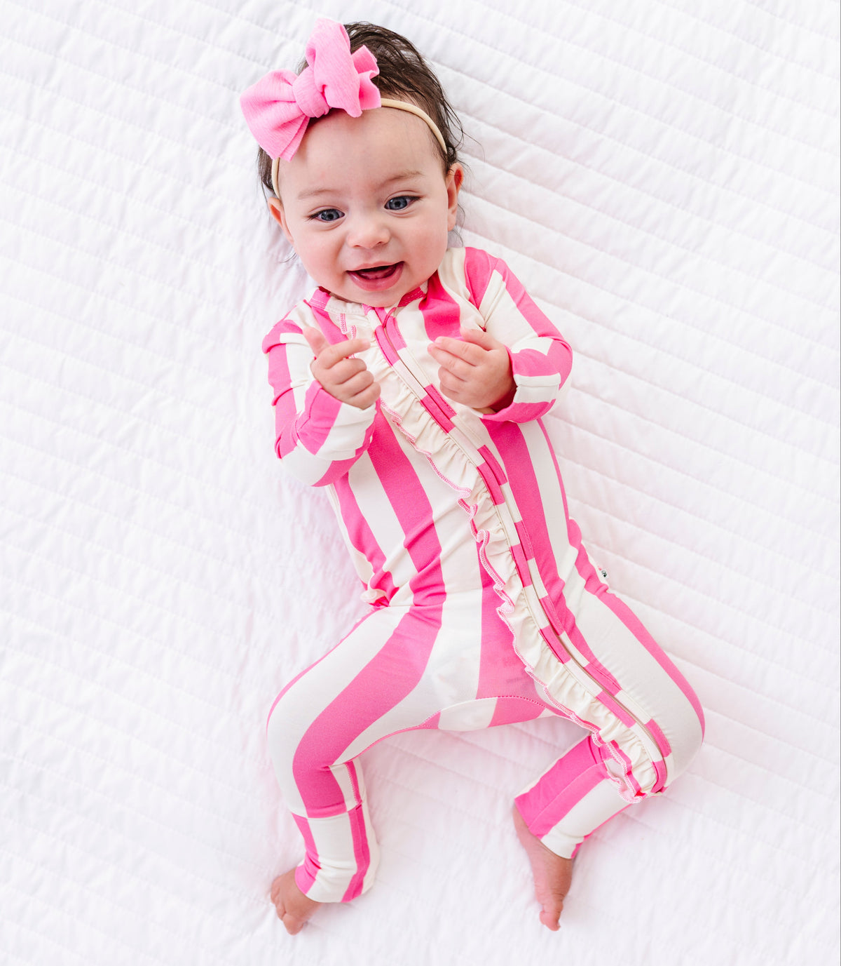 A baby lying on a white quilted blanket, wearing a pink and white striped footless one-piece jumpsuit with ruffle trim and a matching pink bow headband.