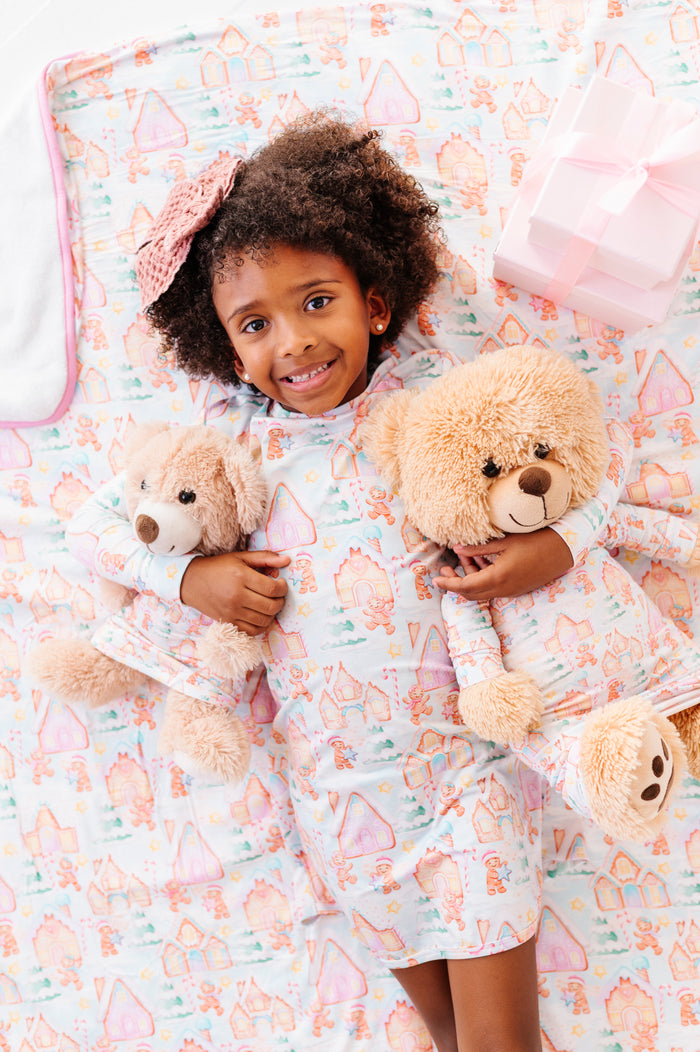 A little girl wearing a bamboo gown featuring a Christmas gingerbread-village themed print, holding two teddy bears wearing matching gingerbread village gowns. They're laying on a plush bamboo blanket