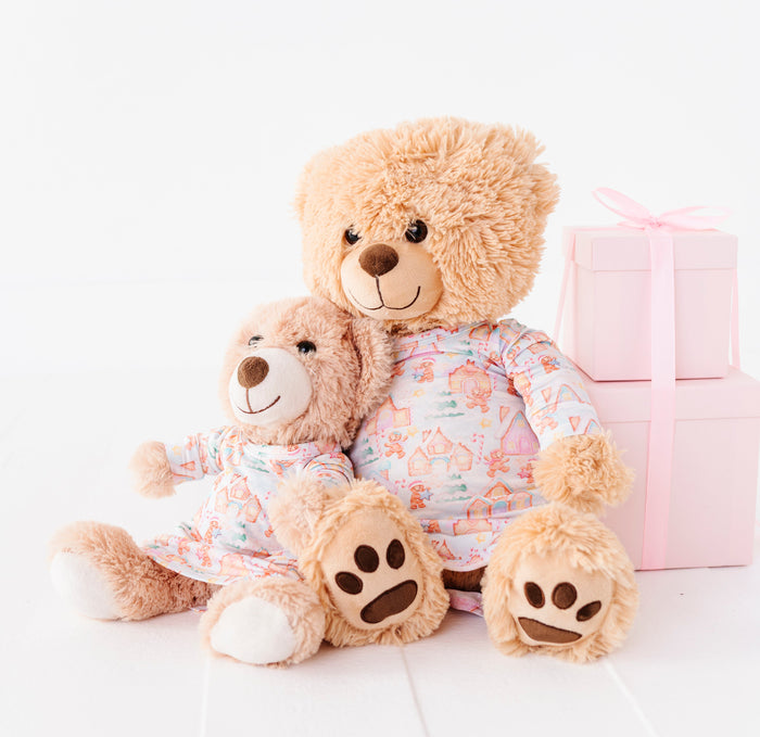 Two teddy bears resting against a pile of pink presents wearing matching Christmas gingerbread village-themed bamboo gowns