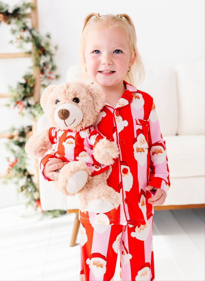 A young girl with blonde pigtails stands indoors wearing red Santa-themed pajamas and holding a beige teddy bear that matches her pajama print. The room has a white sofa, a decorated Christmas tree, and bright natural light.