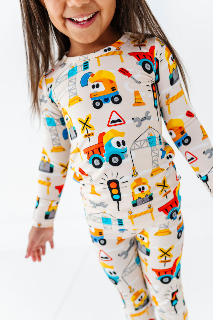 A young child wearing a long-sleeve one-piece pajama top and matching pants with a colorful construction vehicle print on a cream background, photographed against a clean white studio background.