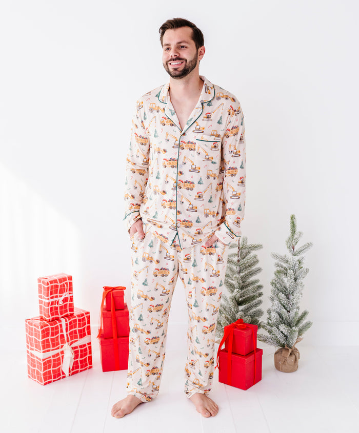A man wearing a long-sleeve bamboo pajama set with a Christmas tree and snowman print stands barefoot in a bright white room, flanked by red wrapped gift boxes and small artificial trees.