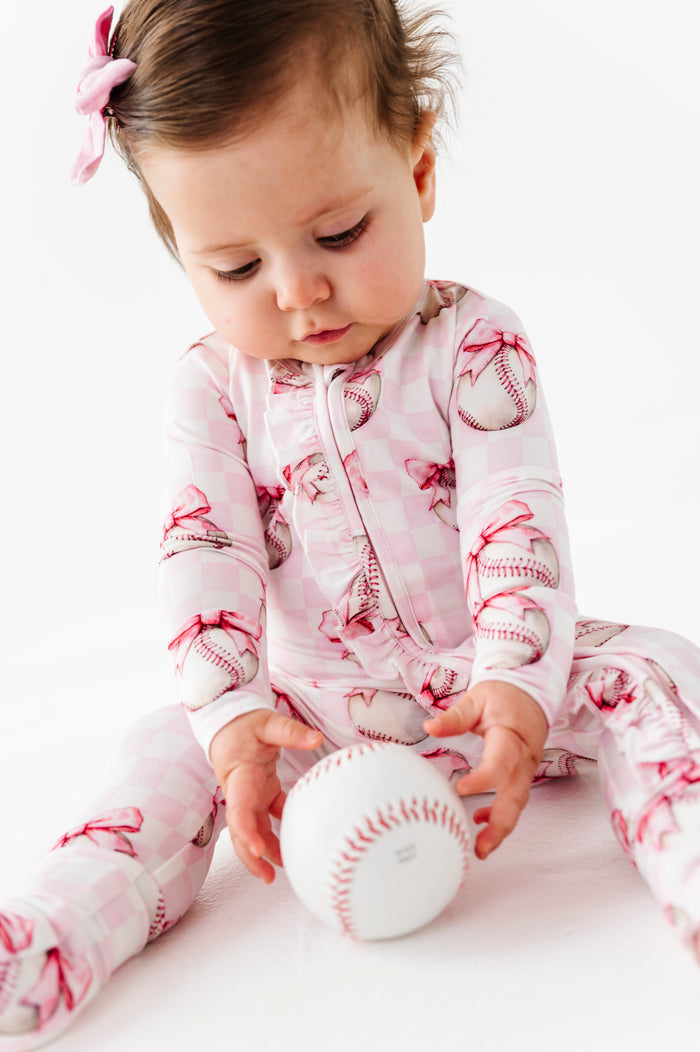 Baby in a baseball-themed bamboo romper playing with a real baseball on a white background