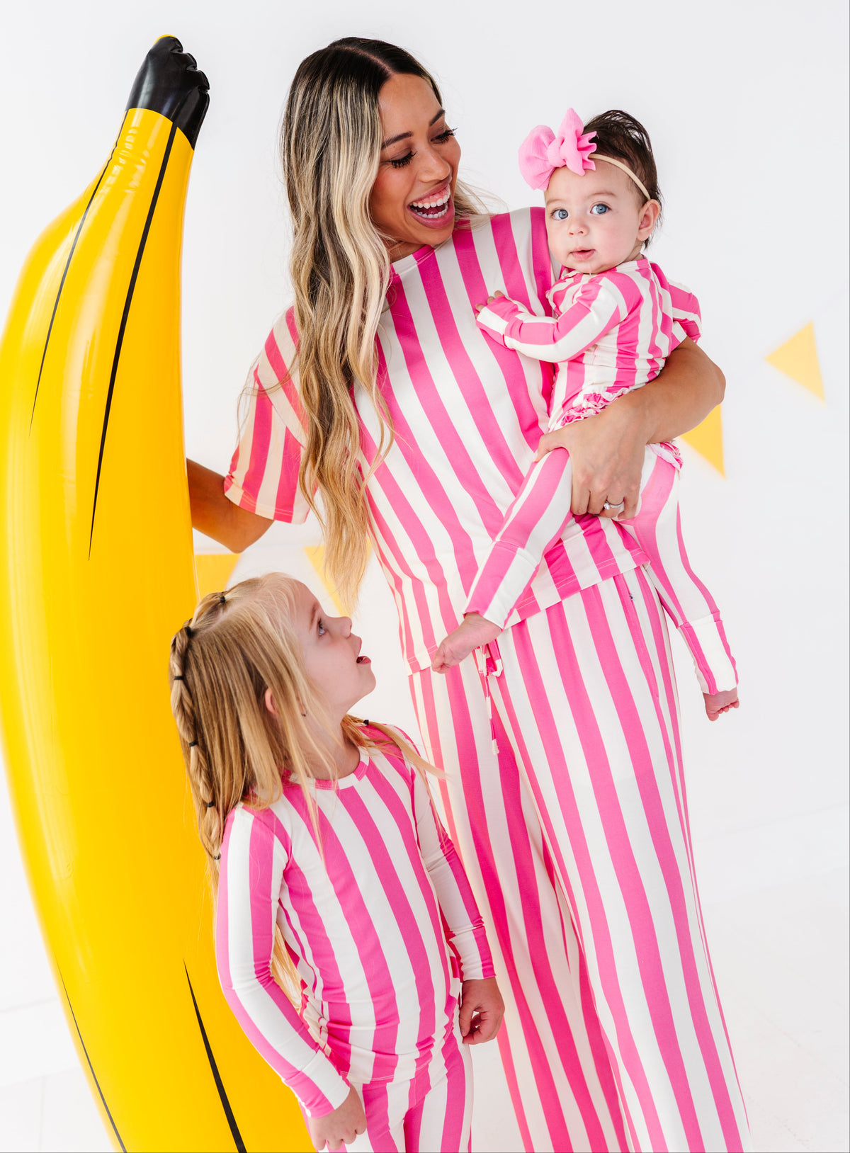 A woman and two children wearing matching pink and white striped one-piece outfits pose with a giant yellow inflatable banana prop against a white studio background.
