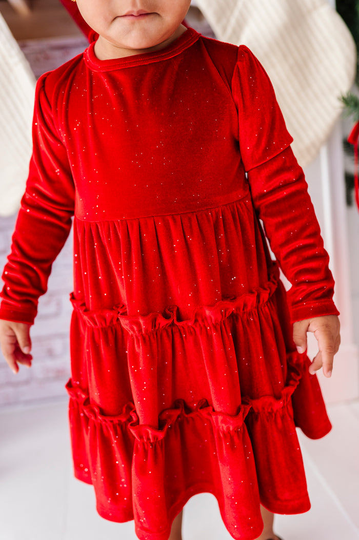 A toddler wearing a bright red long-sleeve dress with a tiered ruffle skirt and subtle glitter, standing in a festive indoor setting.