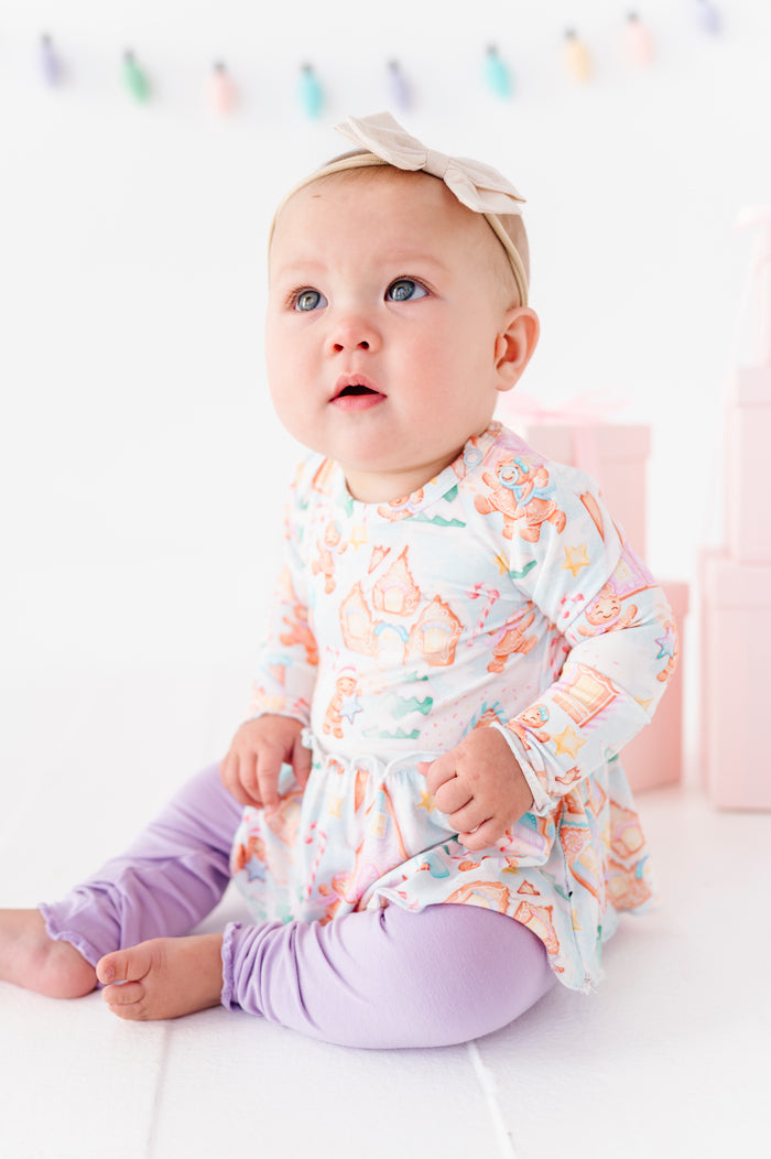 A baby girl wearing a Christmas gingerbread village-themed bamboo peplum with light purple bamboo leggings