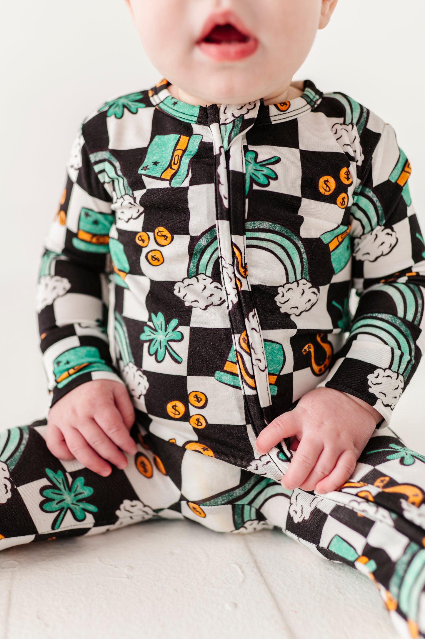 Detail shot of a baby wearing a colorful St. Patrick's Day onesie with top hats, four leaf clovers, gold coins, and rainbows on a white background