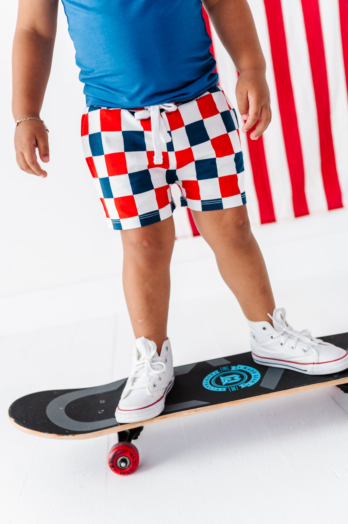 Child on a skateboard wearing red, white, and blue checkered bamboo shorts with an American flag in the background.
