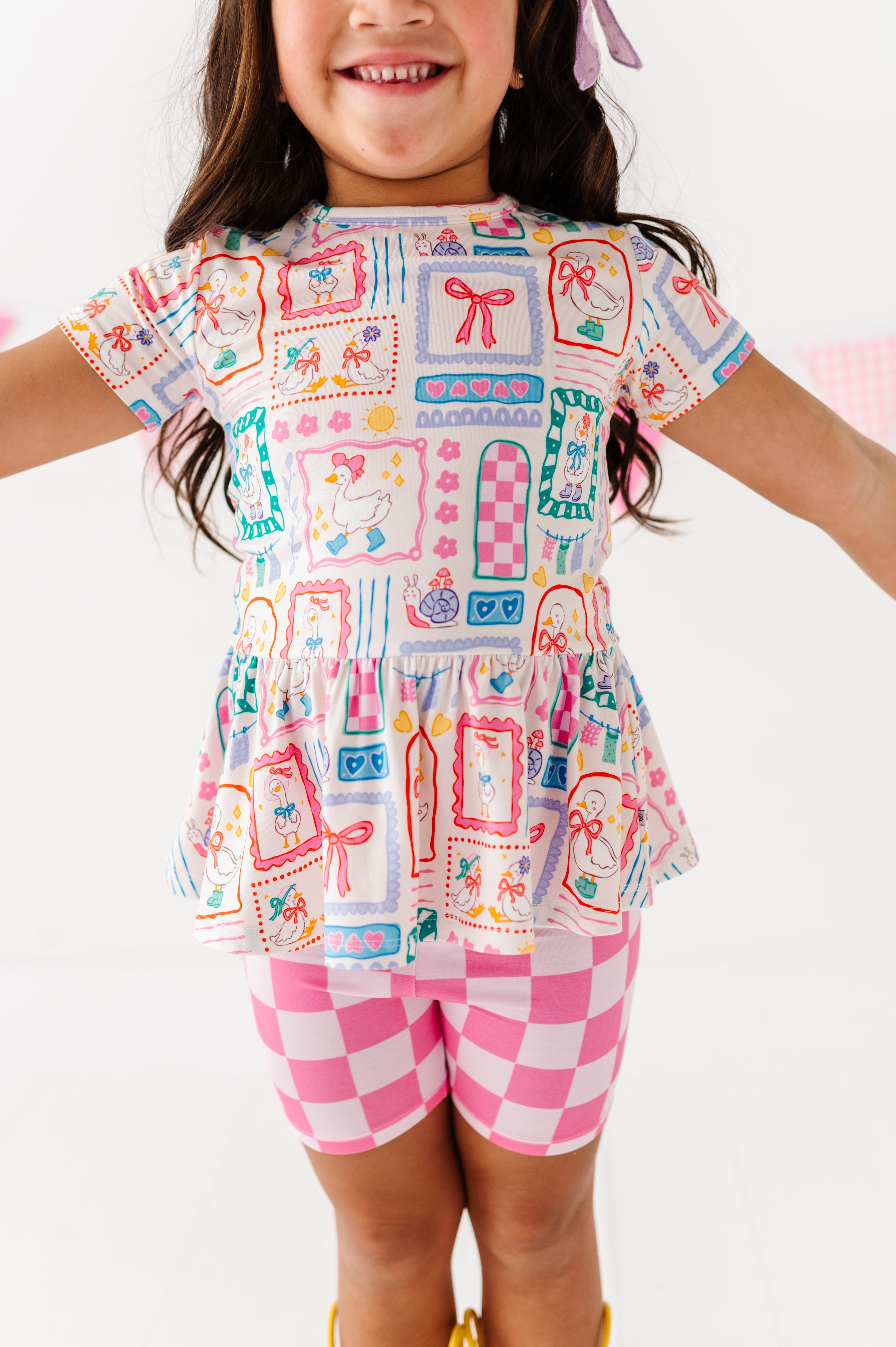 Child wearing a Gilly Goose Bamboo/Cotton Peplum Top and checkered bamboo biker shorts against a white background