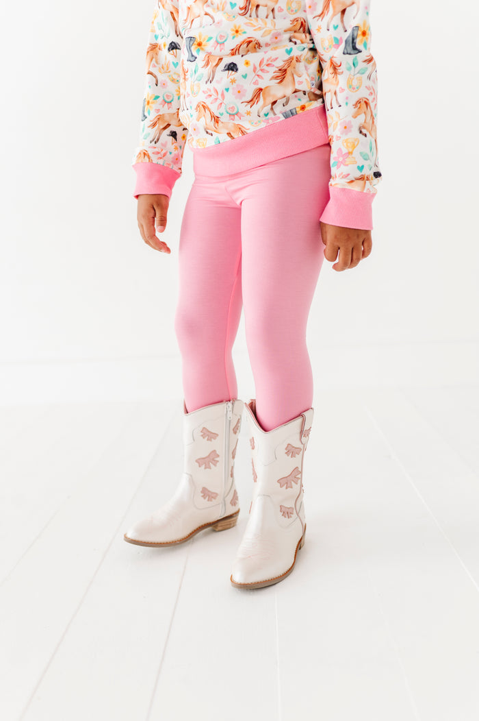 Child wearing pink leggings, horse top, and white boots on a white background