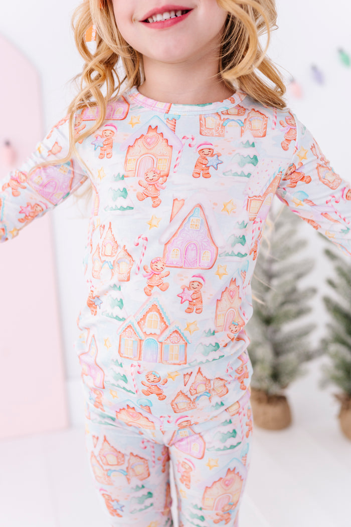 A young girl wearing a long-sleeve bamboo pajama set with a colorful gingerbread house and gingerbread men pattern on a white background, shown in a brightly lit indoor setting.