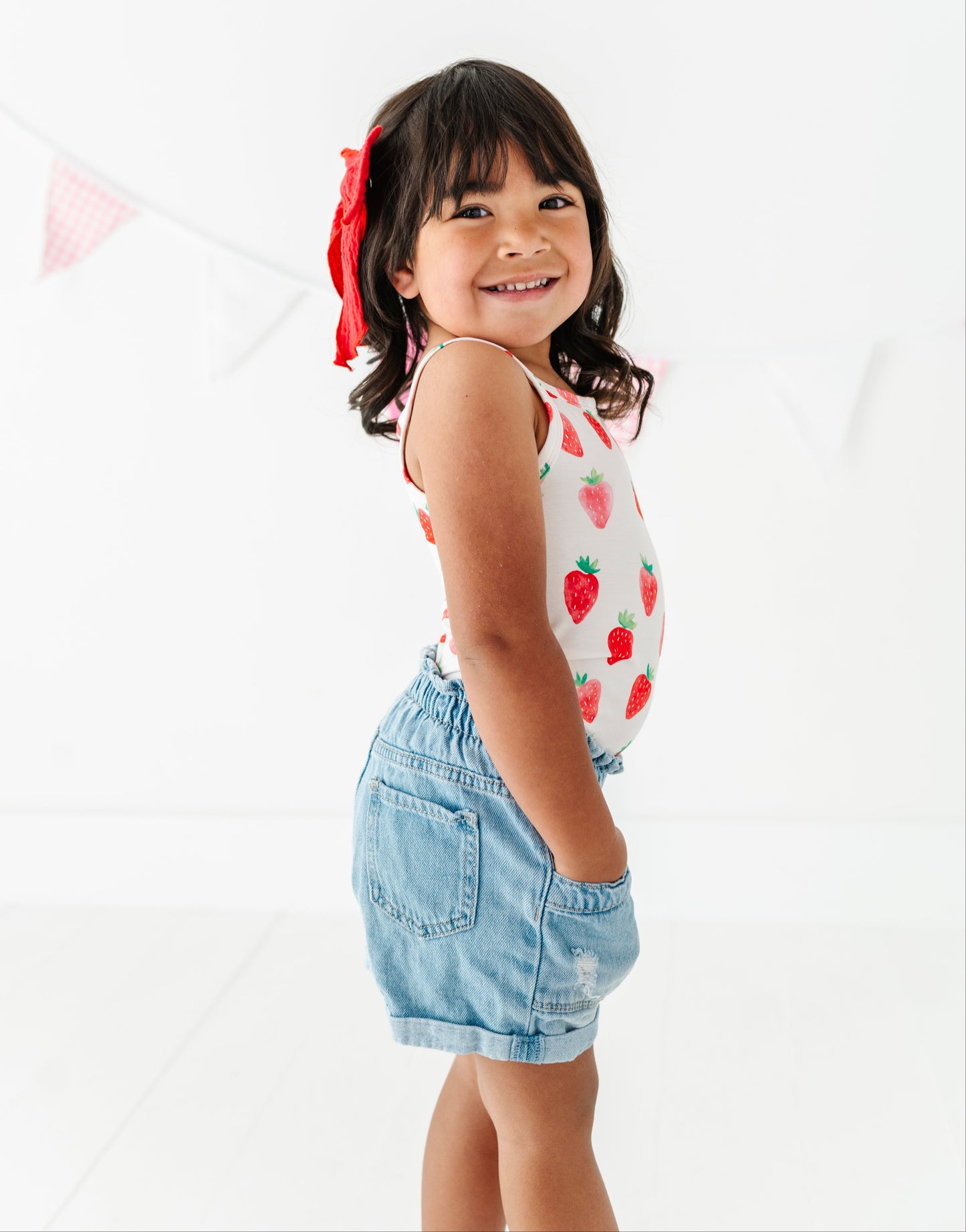 Young girl wearing a tank top with strawberry pattern and denim shorts on a white background