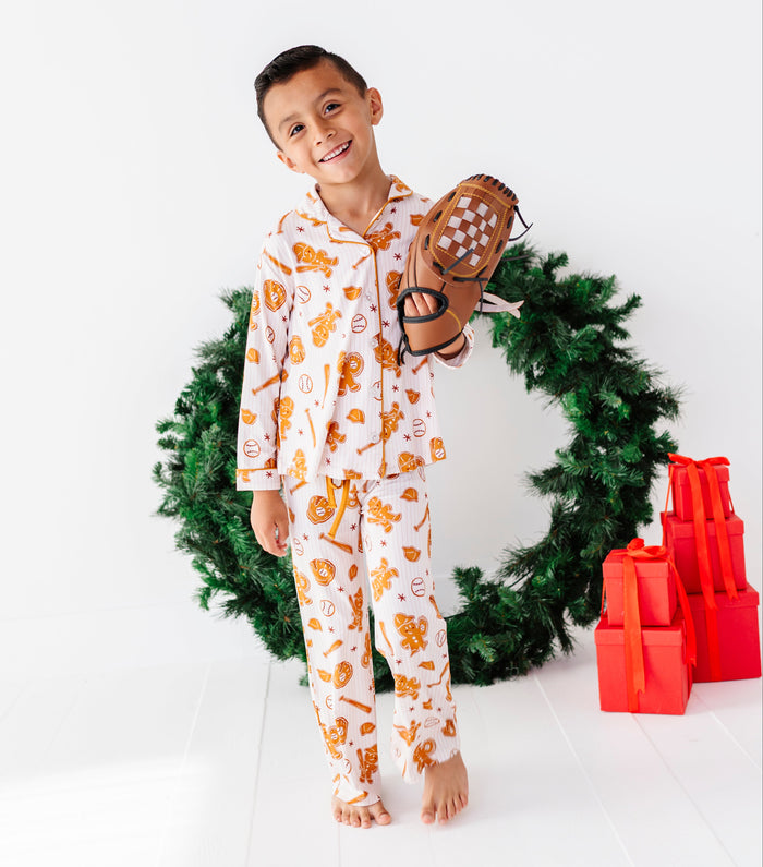 A young boy smiling and holding a baseball glove, wearing a white long-sleeve pajama set with an orange gingerbread and cookie pattern. He stands barefoot in front of a green Christmas wreath with red wrapped presents on a white background.