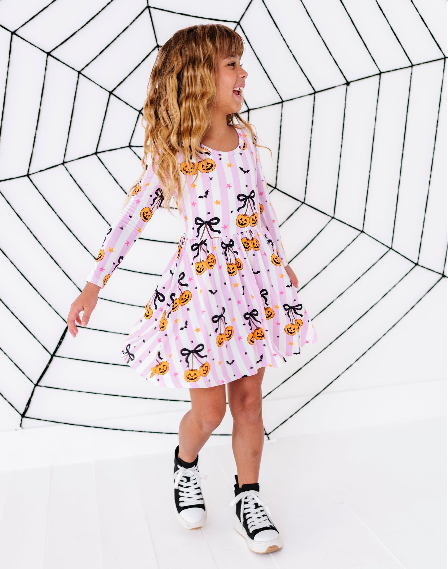 A young girl with blonde curly hair wearing a pink long-sleeve dress with a pumpkin and bow pattern, standing in front of a large black spiderweb prop on a white studio background.