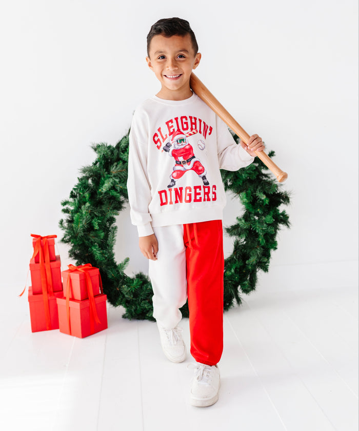 A young boy stands in a studio setting wearing a white long-sleeve sweatshirt with a red and green Christmas graphic that reads 'Sleighin' Dingers' and matching white sweatpants. He is holding a wooden baseball bat over his shoulder, with a green wreath and red gift boxes behind him on a white background.